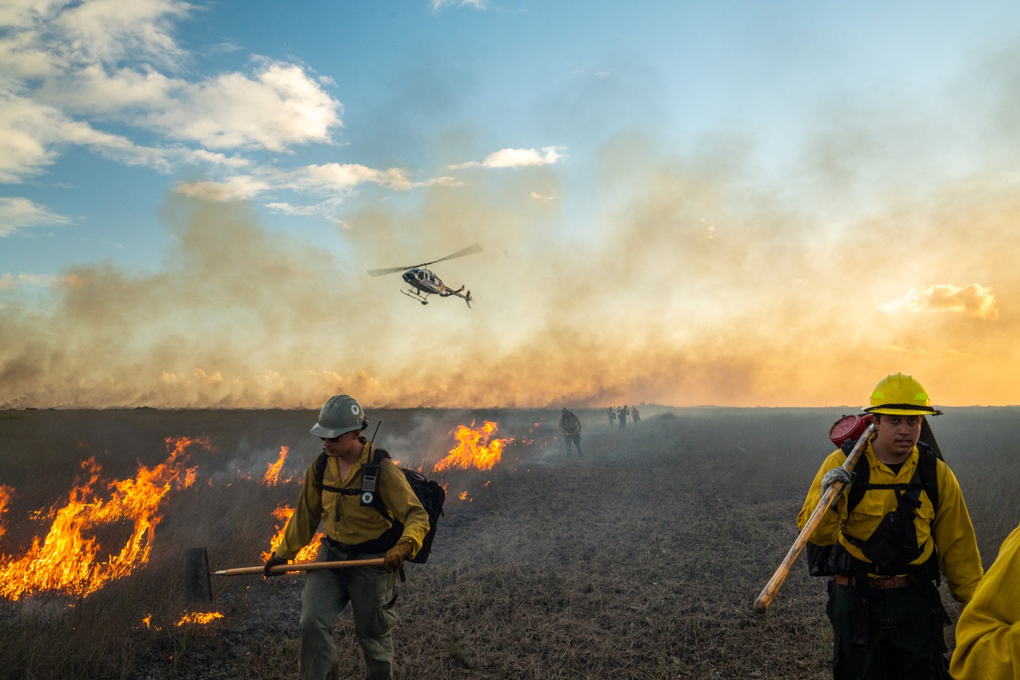 Prescribed_burn_on_prairie_NPS_photo_by_Ian_Wilson Firefighters walk near a burning field with a helicopter flying overhead.