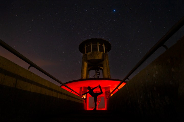 Person in yoga pose under tower with red light, starry sky above.