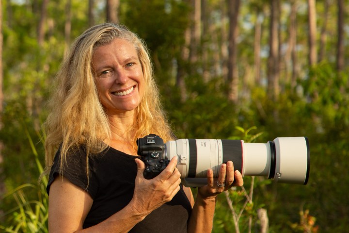 Woman smiling and holding a camera with a long lens in a forest.