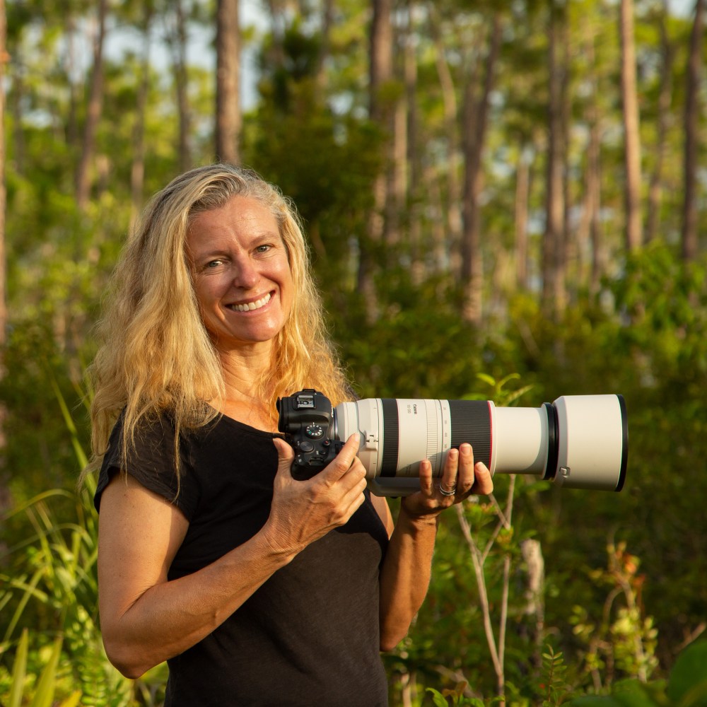 Woman smiling and holding a camera with a long lens in a forest.