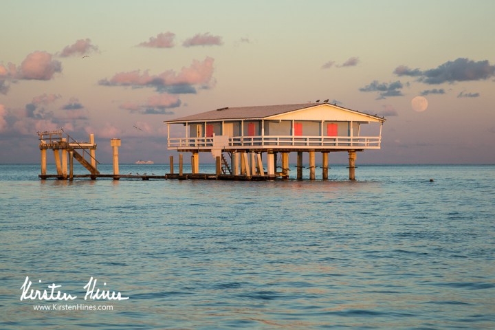 Stilt house over water at sunset, with the moon visible in the sky.