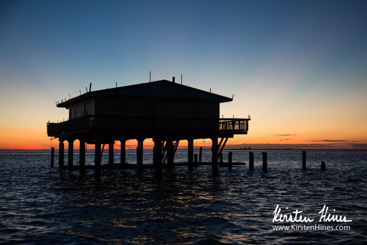 Silhouetted house on stilts over water at sunset with orange sky.